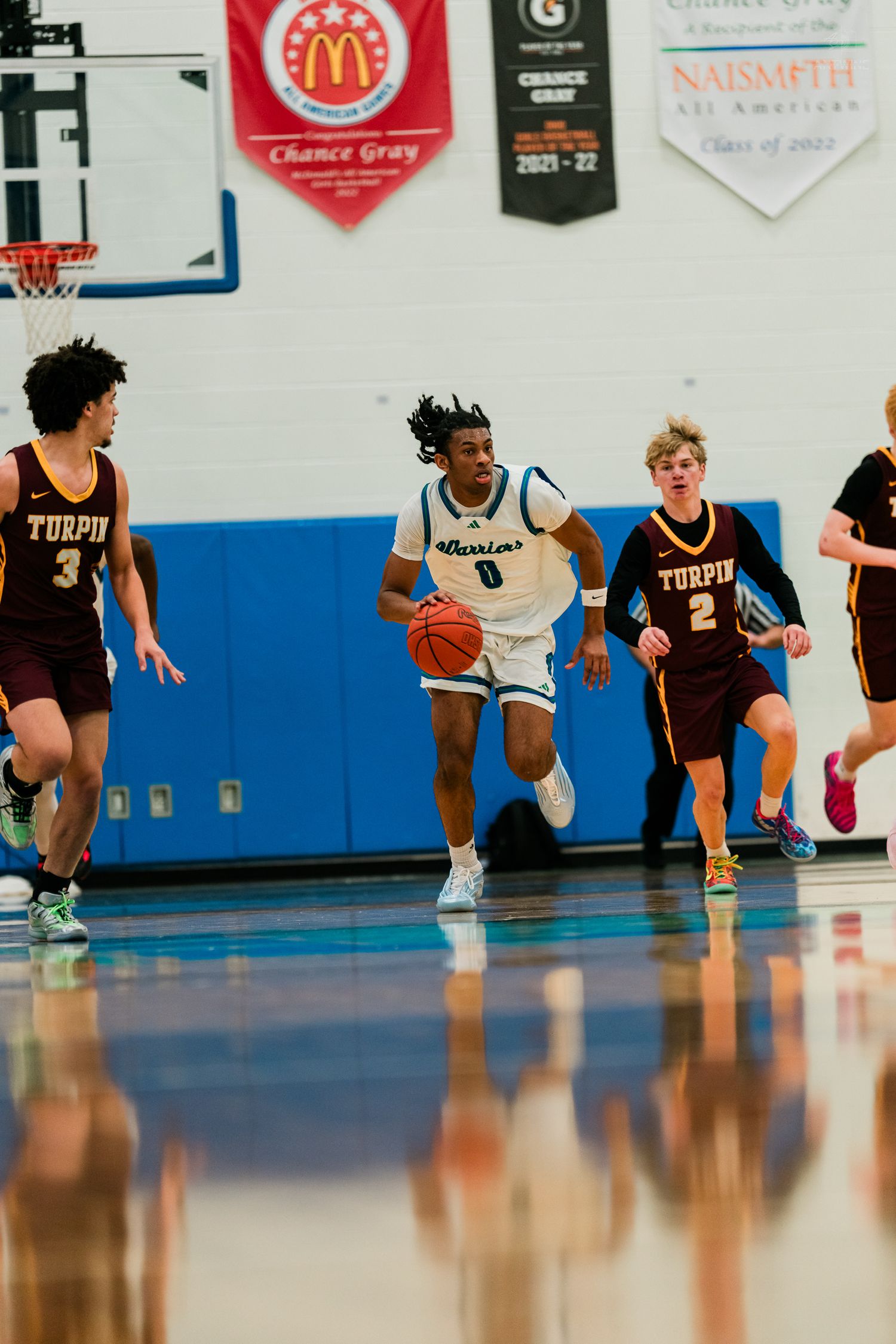 High school basketball player #0 in white Warriors uniform dribbles past Turpin defenders on blue court.