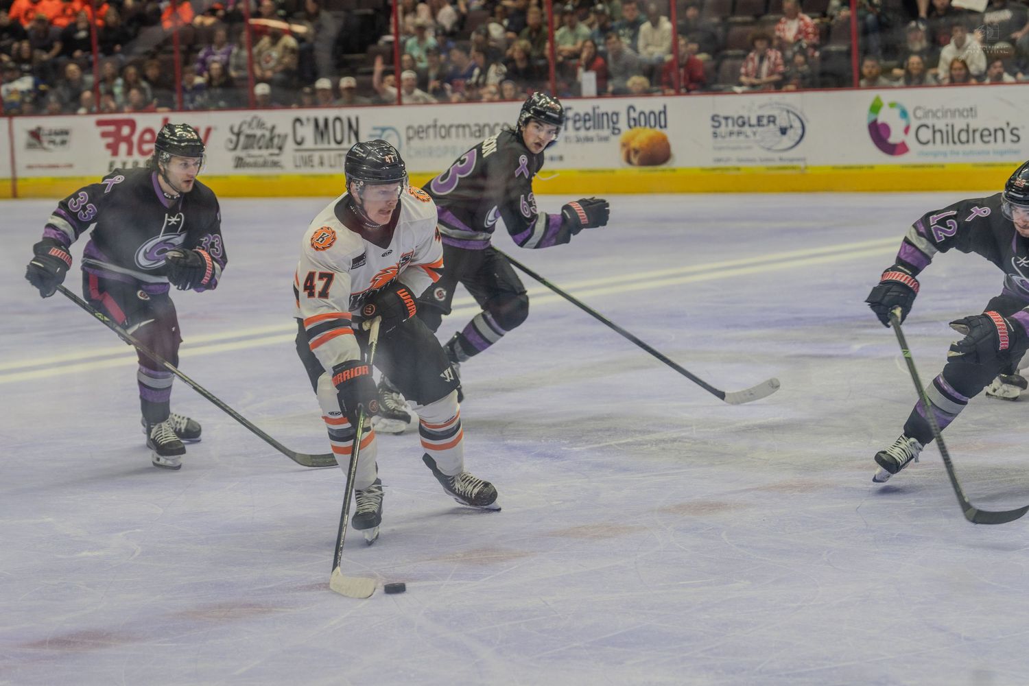 Hockey player #47 in orange jersey controls puck while three defenders in dark uniforms pursue during indoor ice hockey game.