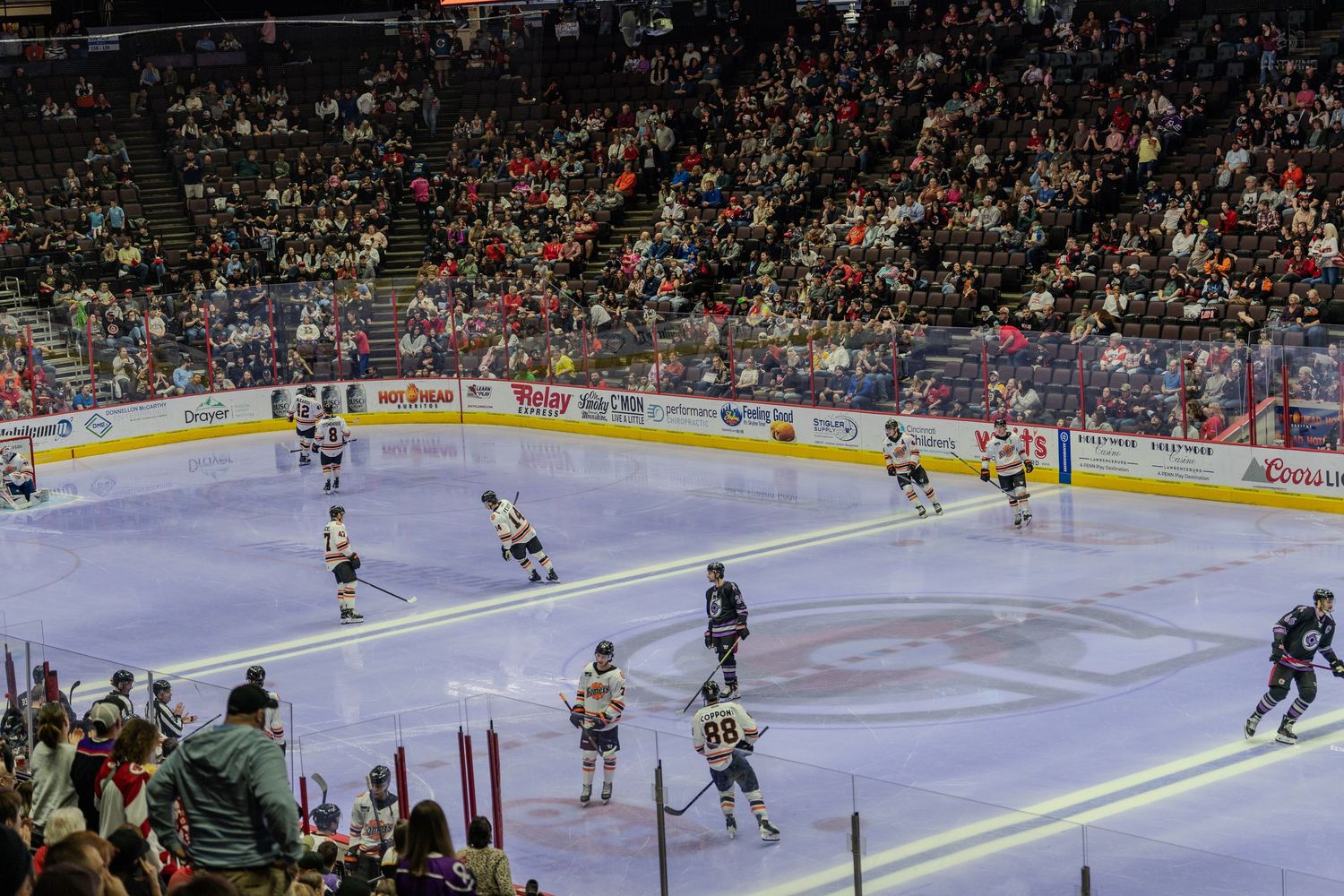 Hockey players skating on the ice during a game at a well-attended indoor arena with a full crowd.