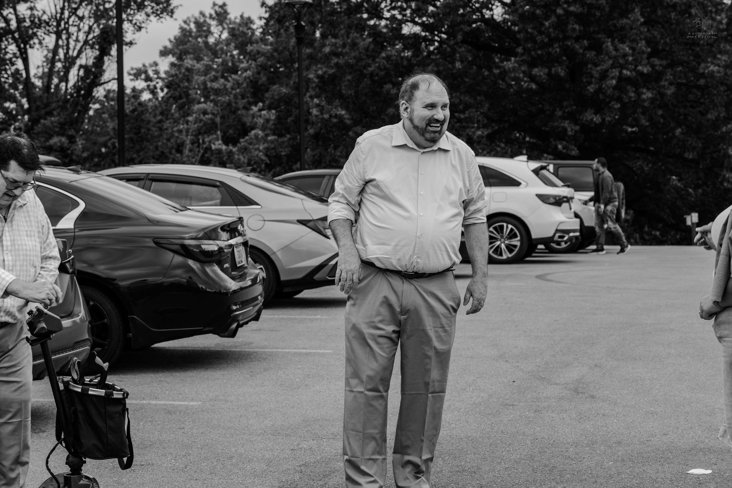 Black and white photo of a man in a button-up shirt smiling in a parking lot with cars.