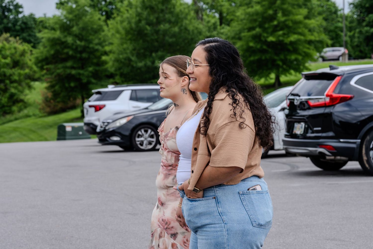 Two women smiling and walking together in a parking lot surrounded by cars and green trees.