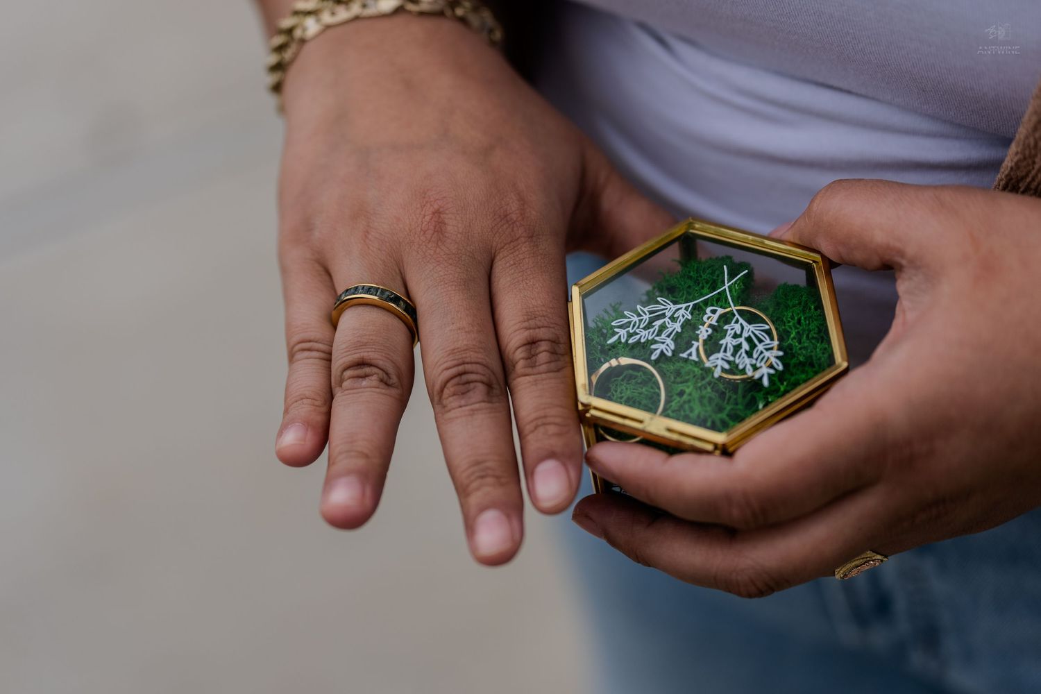 Hands wearing a gold ring holding a hexagonal glass box with green moss and jewelry.
