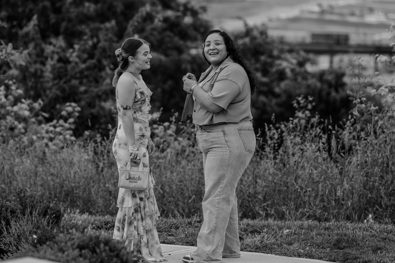 Two women share a lighthearted moment outdoors, one in floral dress and one in casual wear, black and white.