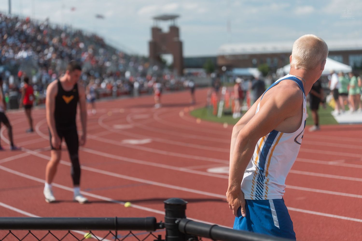 Athlete in blue track uniform leans on railing watching competitors on red outdoor track during a field meet.