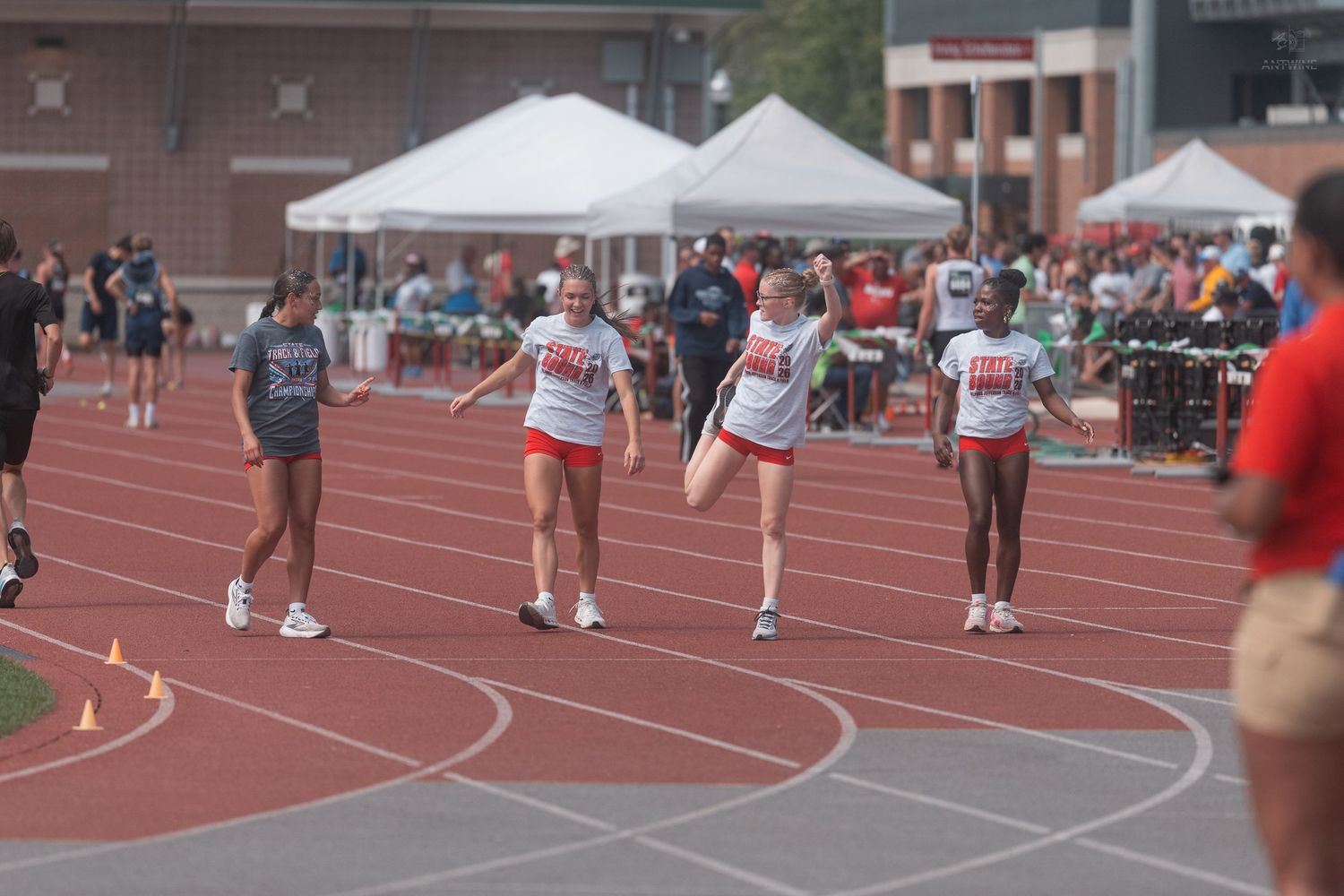 Female athletes in red and white uniforms warm up on a red track at an outdoor track meet.