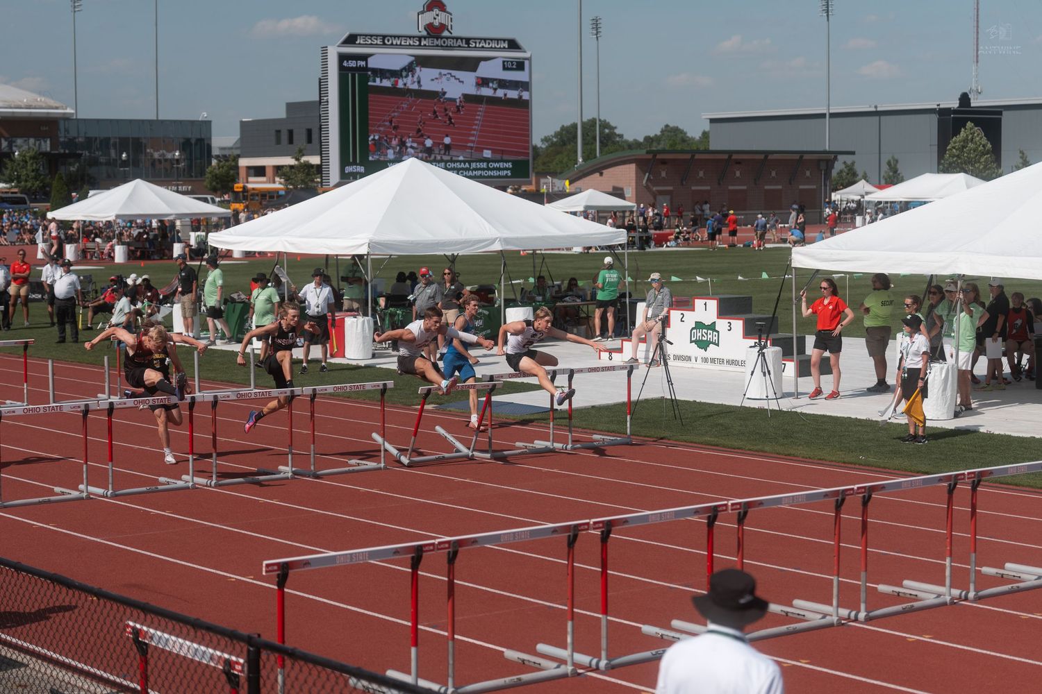 Track athletes clearing hurdles near finish line at outdoor high school championship meet with scoreboard visible.