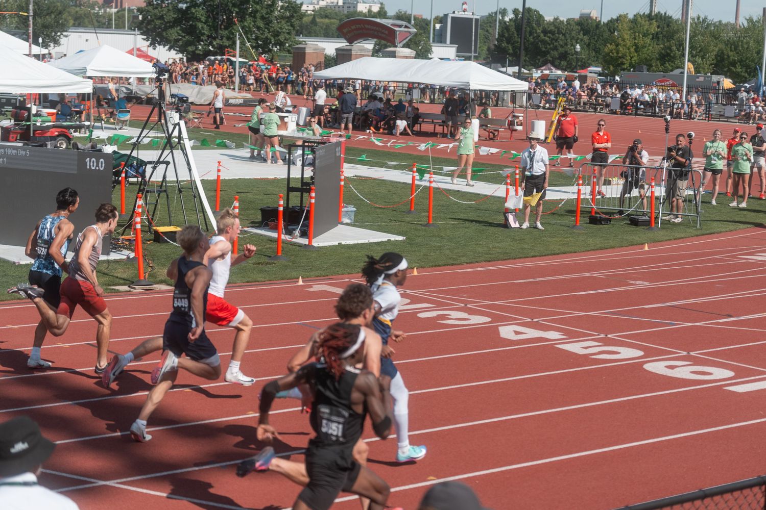 Competitive runners race on a red track during an outdoor track and field event with spectators in the background.