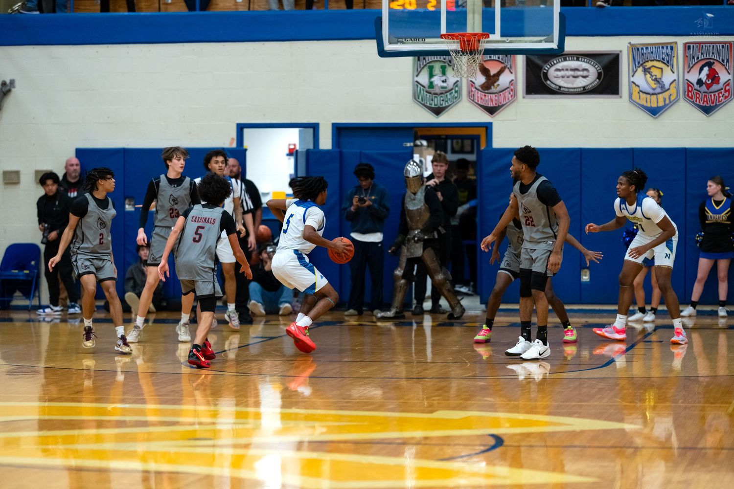 High school basketball players compete on a yellow-floored gymnasium court with spectators watching from the sidelines.