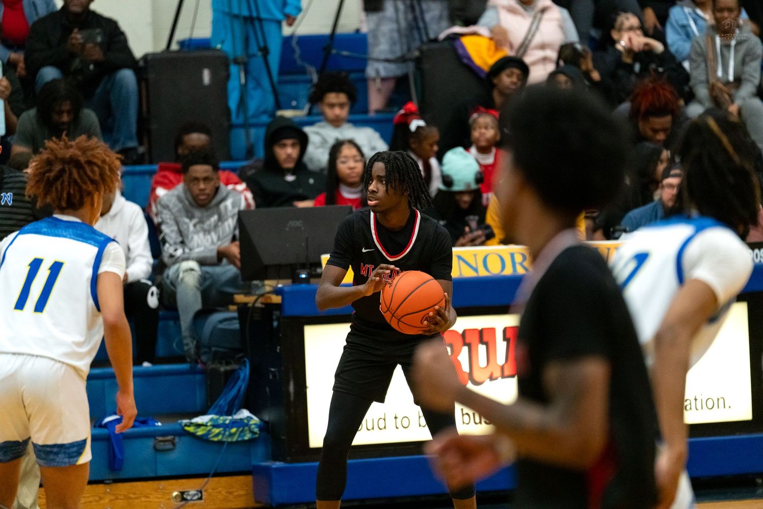 Player in dark jersey holds basketball near scorer's table during a high school basketball game.