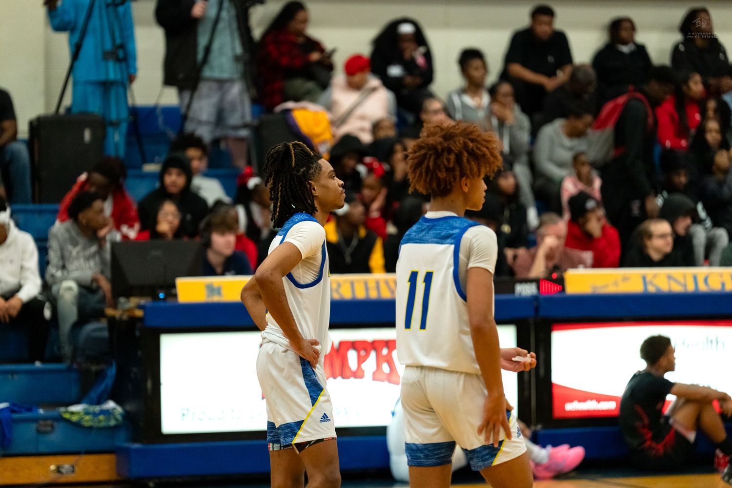 Two high school basketball players in blue uniforms number 11 chat during a timeout on the court.