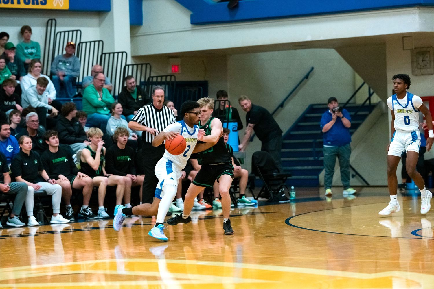 High school basketball player in blue uniform dribbles past green-uniformed defender with teammate number 0 in background.