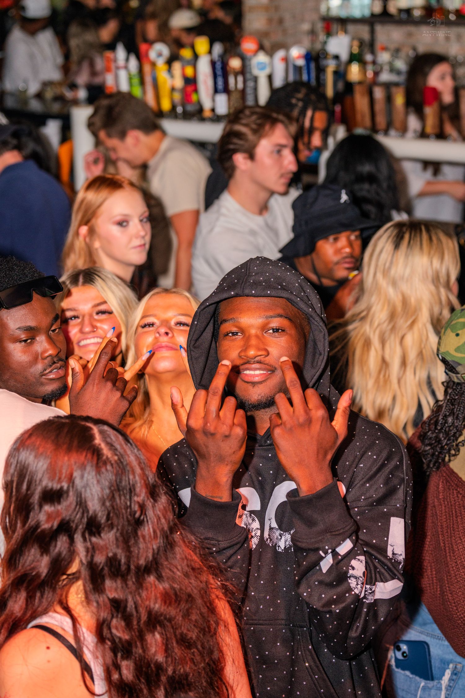 Smiling man in polka dot hoodie posing with hand gestures at a crowded bar event.