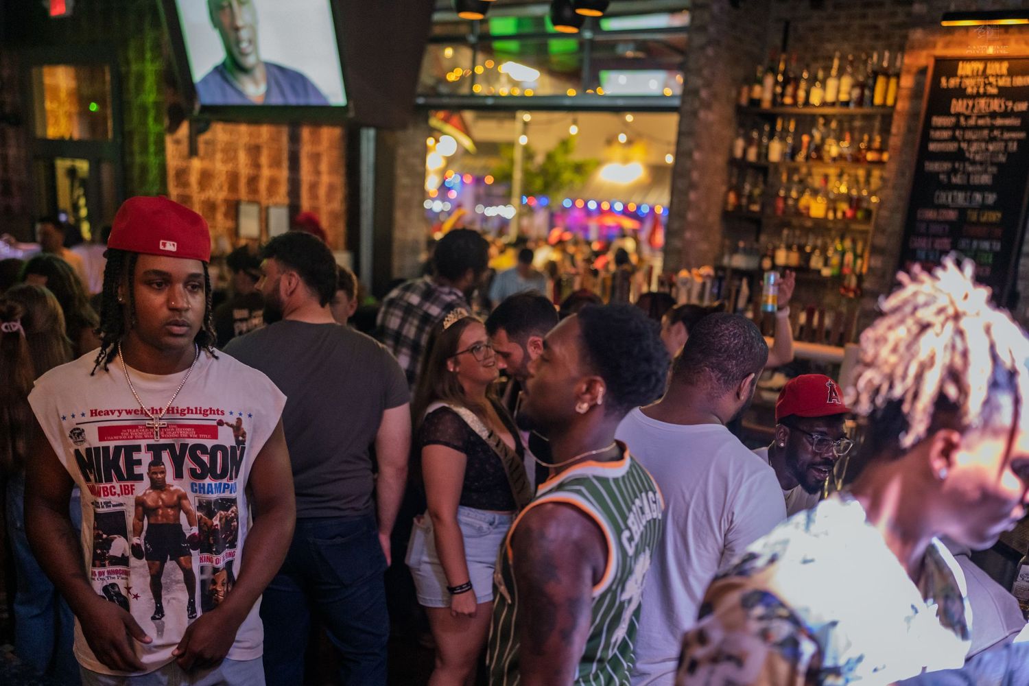 Crowded nightclub bar scene with diverse group of young adults socializing, man in red cap wearing Nike Tyson shirt stands out.