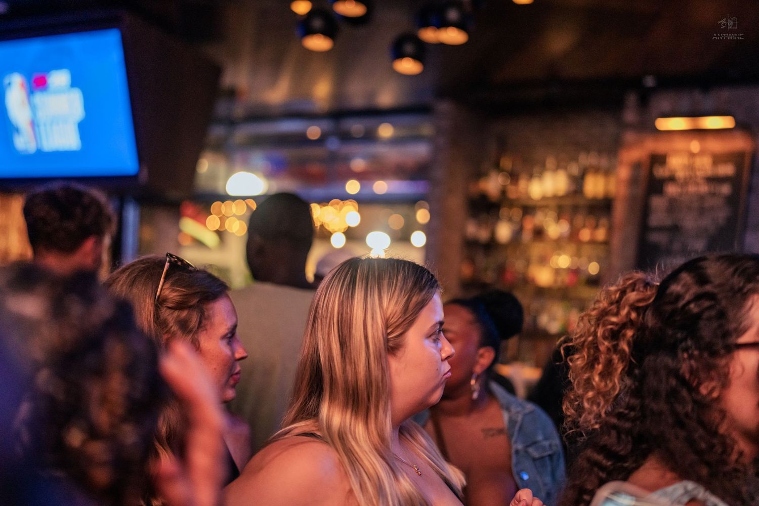 Crowd of people socializing at a dimly lit bar with a TV screen in the background.