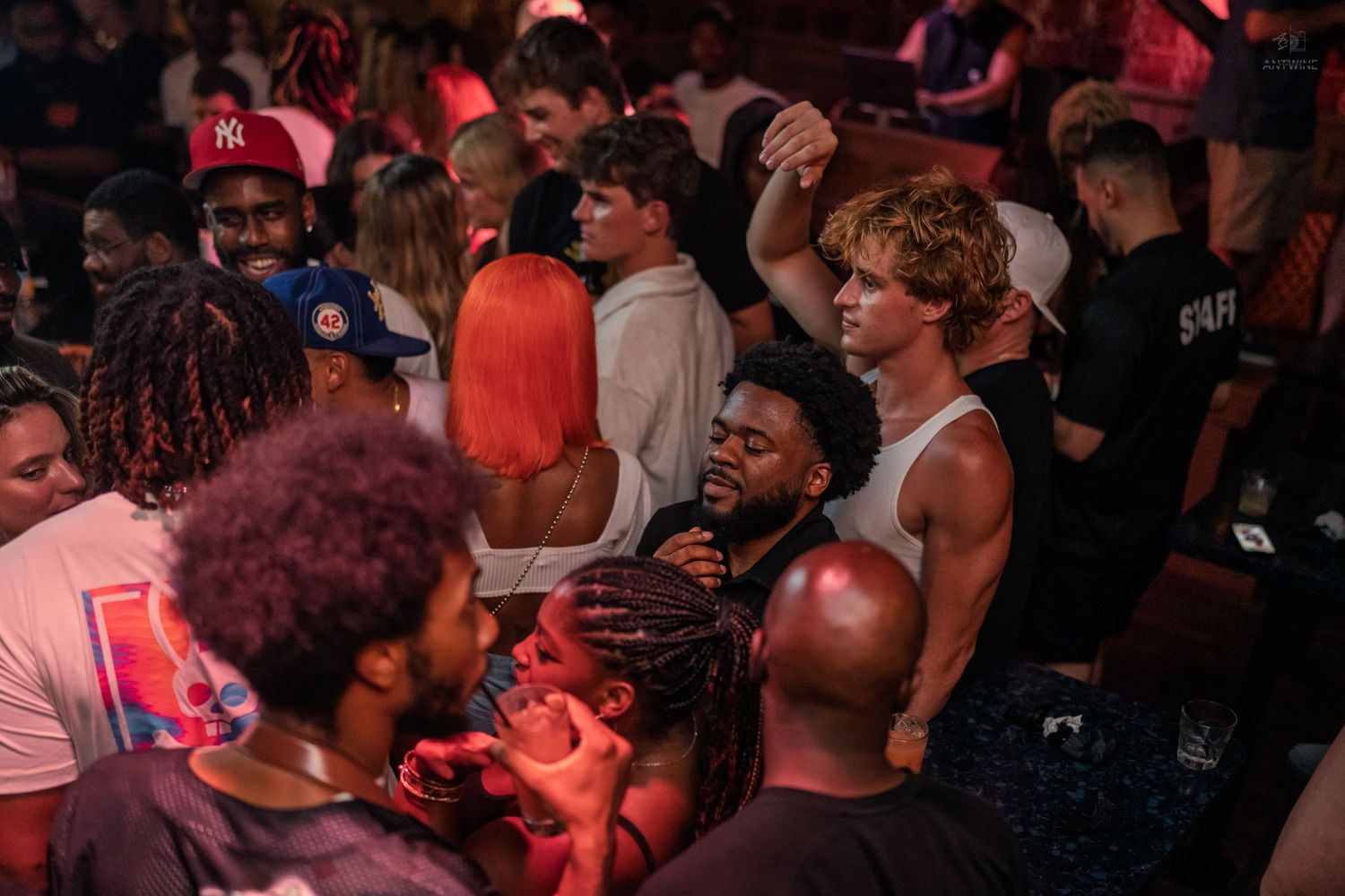 Diverse crowd of people dancing and socializing at a vibrant nightclub party with red ambient lighting.