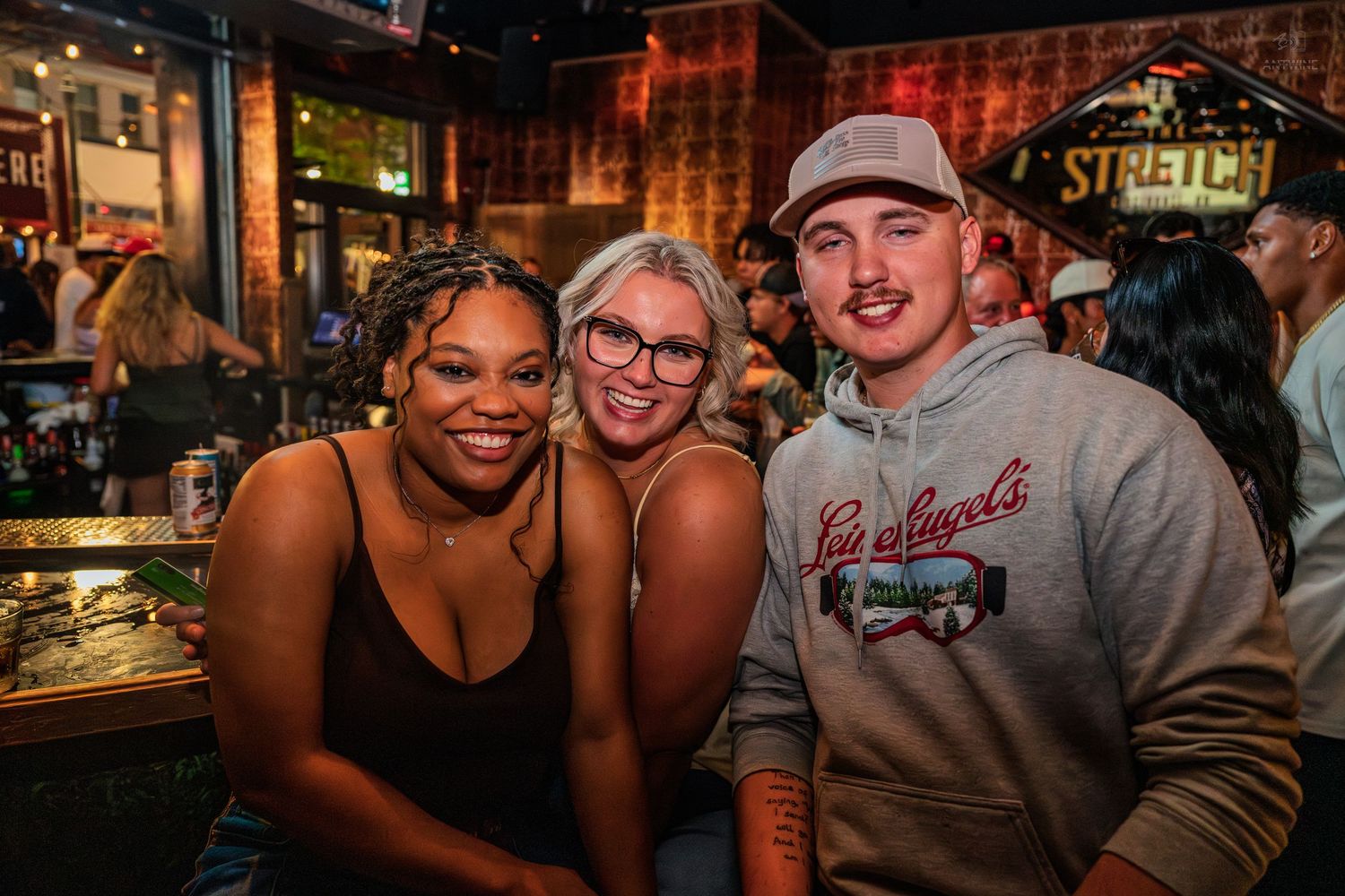 Three friends smiling together at a bar with Stretch branding visible in the background.