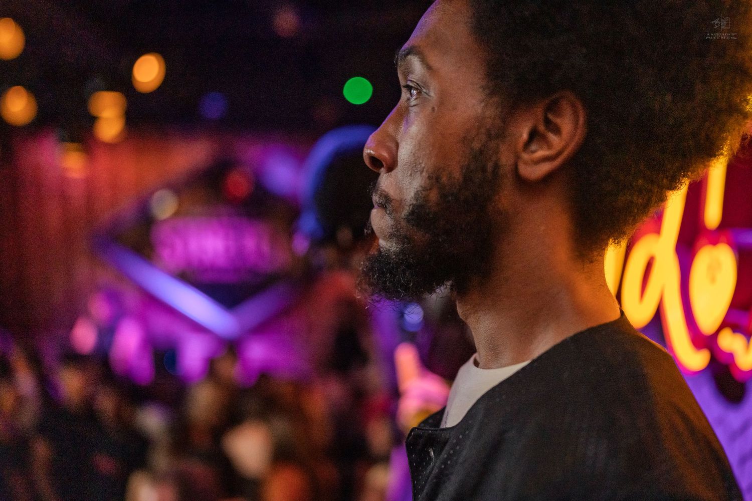 Man with braided hair and a watch gesturing while attending a lively indoor bar event.