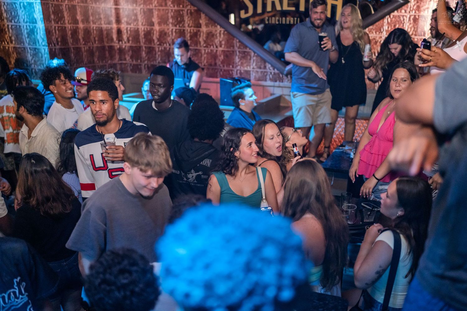 Crowded nightclub scene with diverse group of young adults socializing and dancing under blue lights.