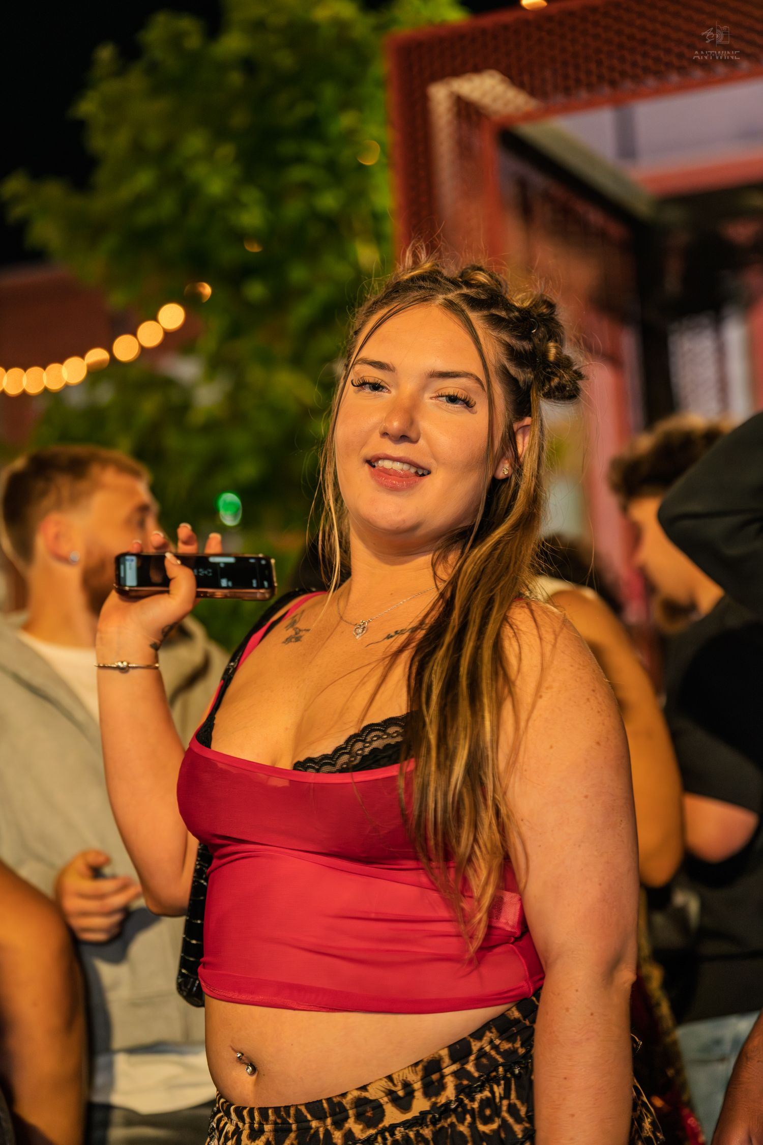 Smiling young woman in red crop top holding a phone at an outdoor night event.