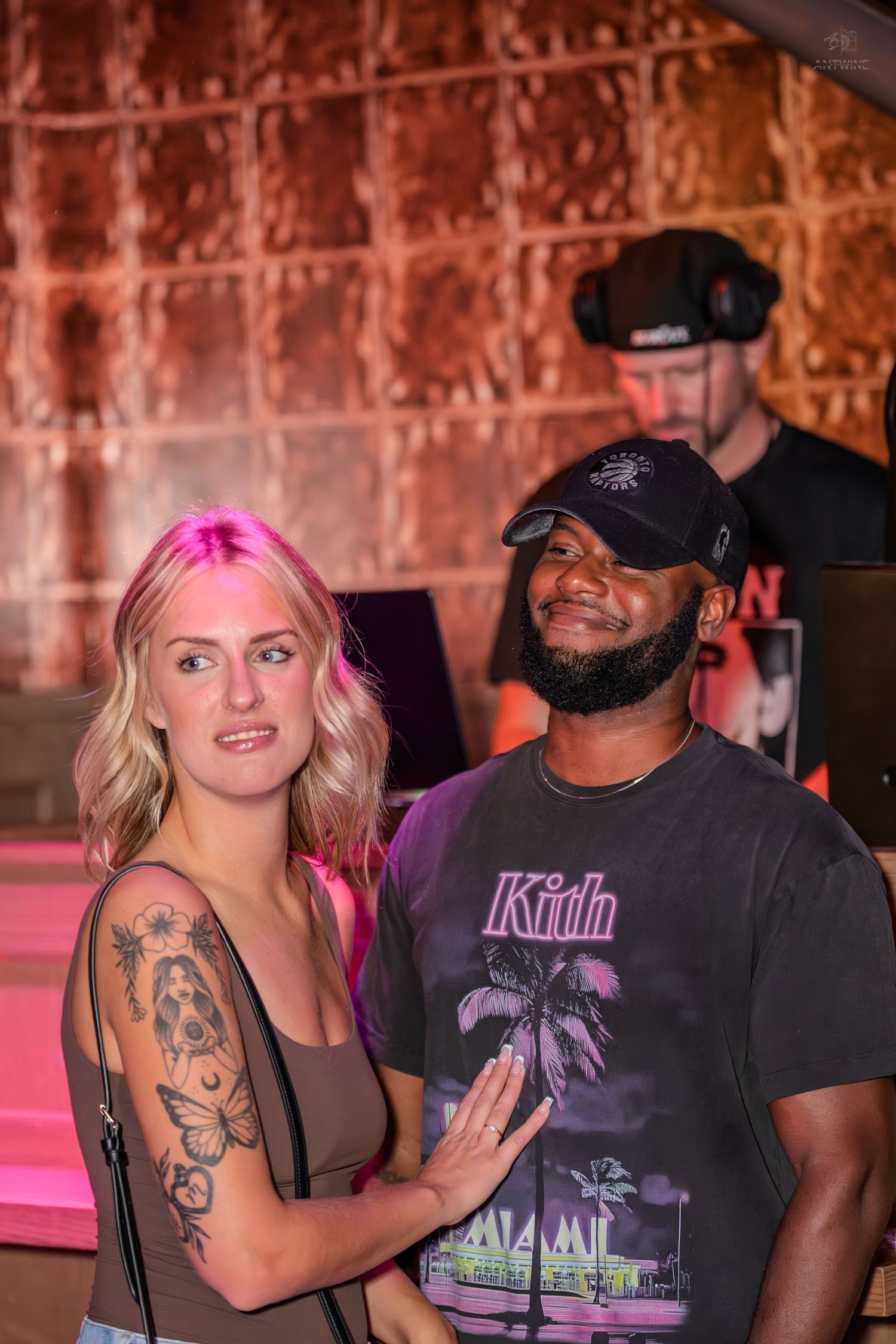A man and woman pose together at a nightclub with a DJ performing behind them.