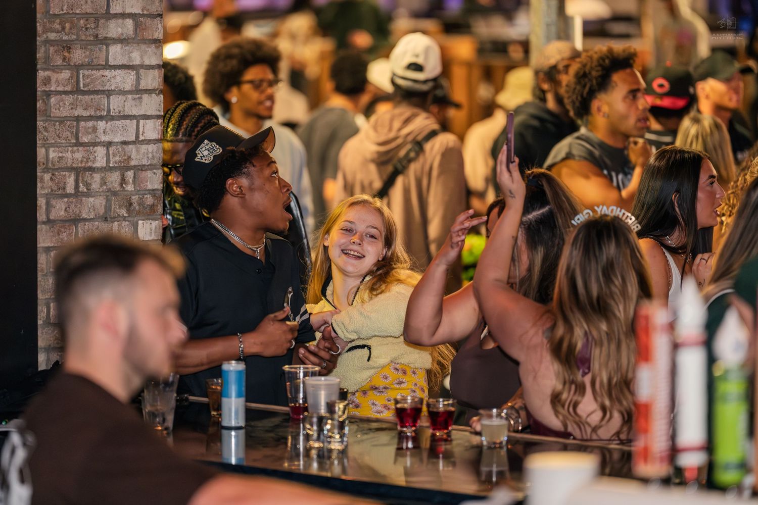 Lively bar scene with diverse crowd of young adults socializing and laughing around a counter with drinks.