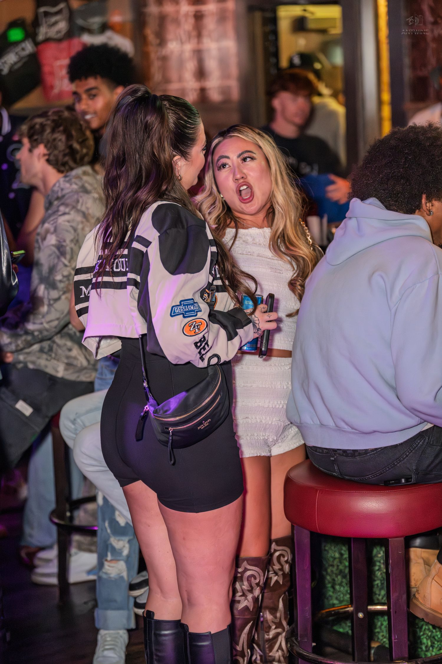Two young women at a lively bar, one wearing a fanny pack, the other reacting with an open-mouthed expression.