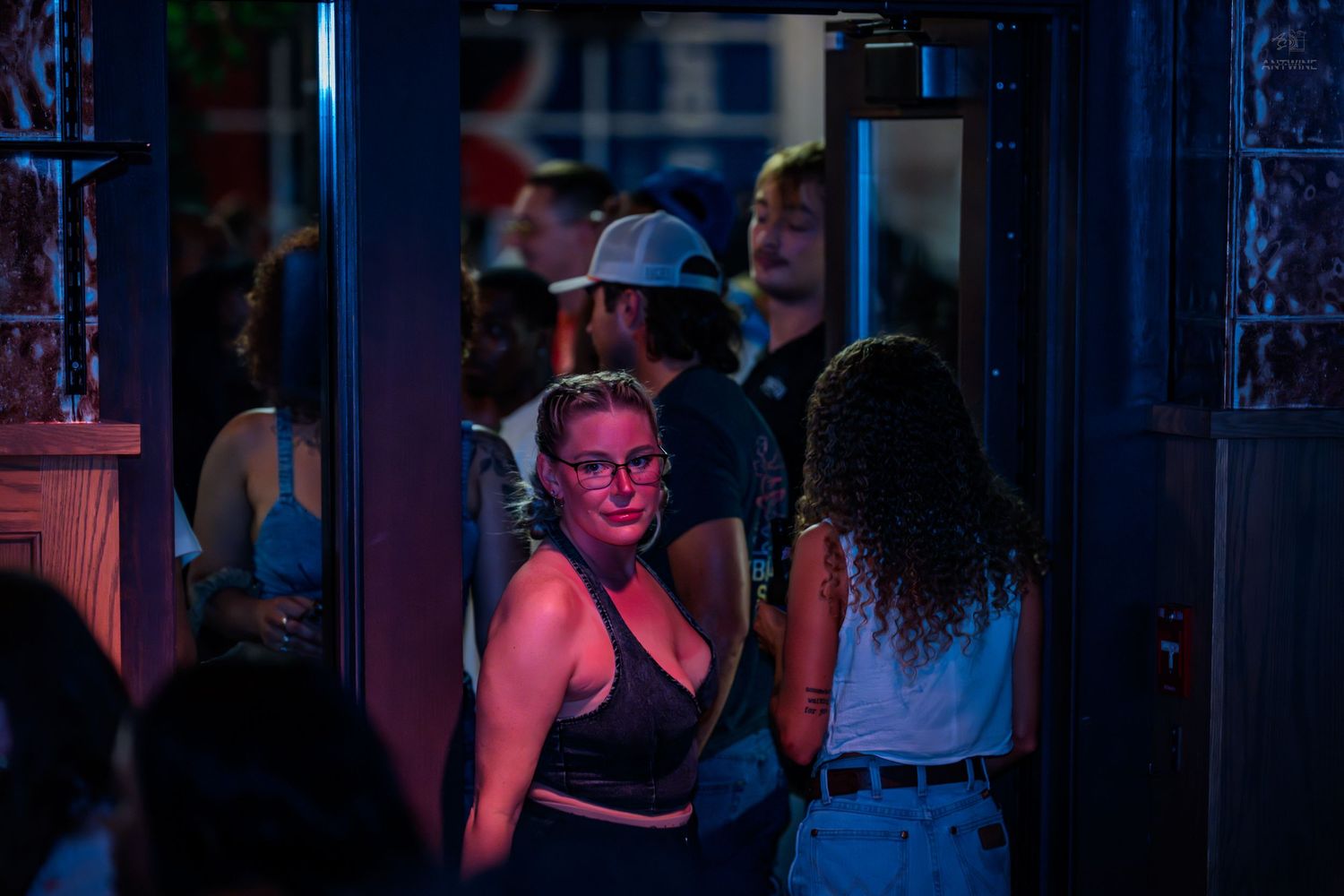 Young woman in glasses smiling near a nightclub entrance with a crowd of people behind her.