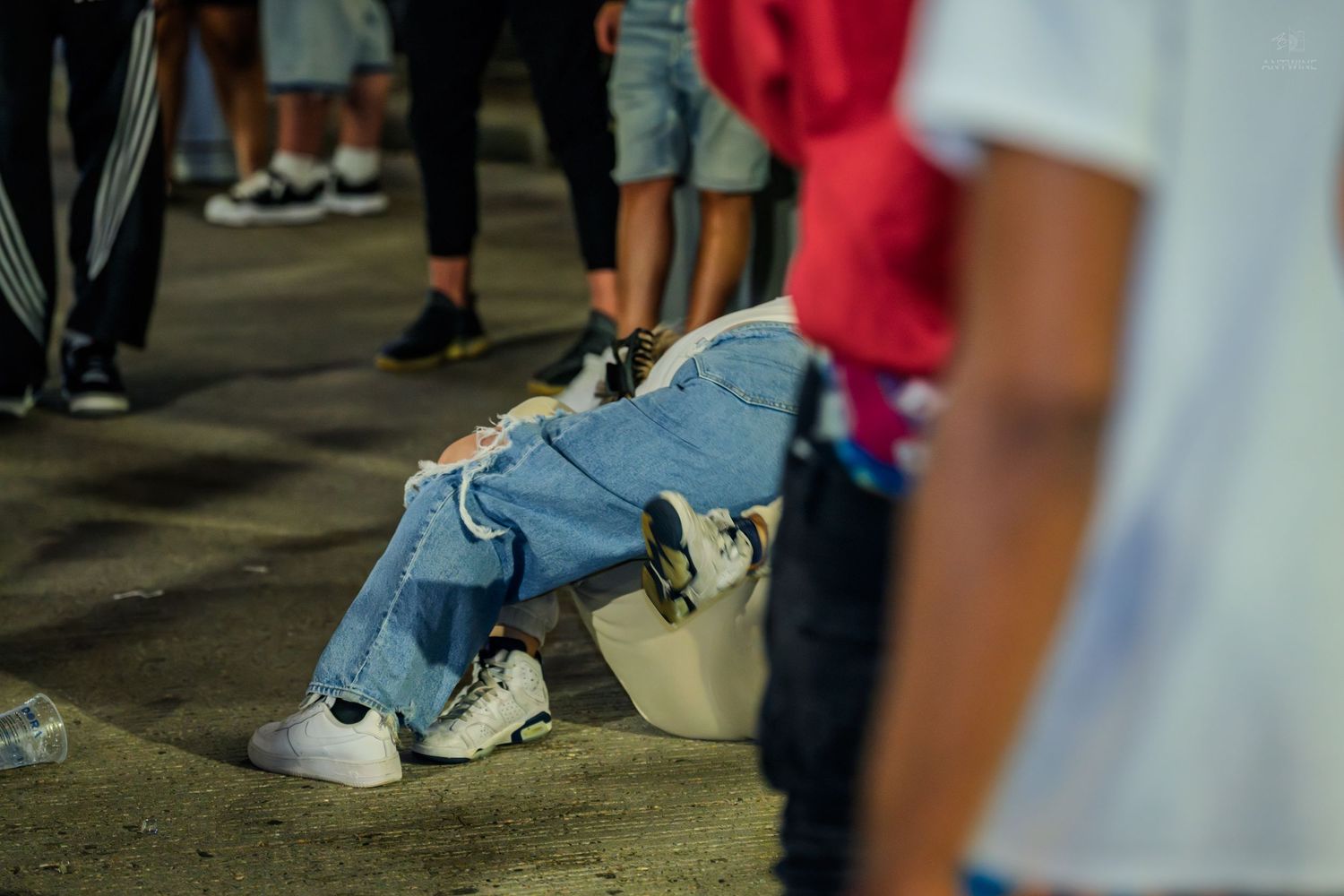 Person crouching low to the ground in ripped jeans and white sneakers at an outdoor event.