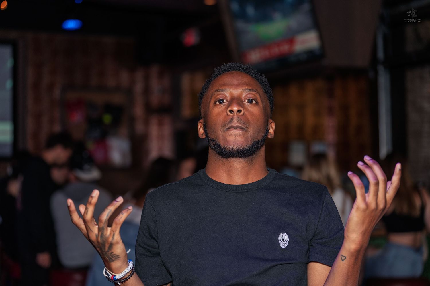 Man in a black t-shirt gesturing with both hands raised in a busy bar setting.