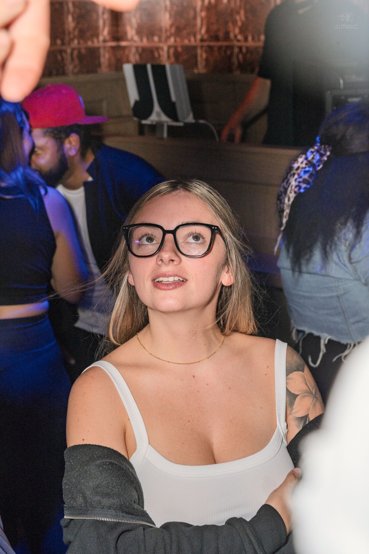 Young blonde woman wearing black glasses and white top smiles at a crowded nightclub party.
