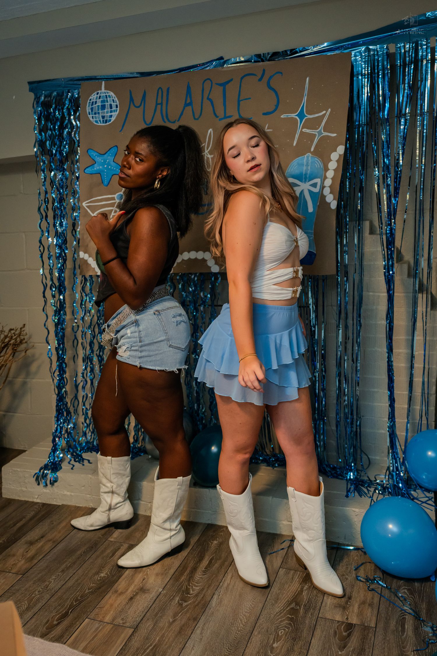 Two women in white cowboy boots pose back-to-back at Malarie's blue 21st birthday party.