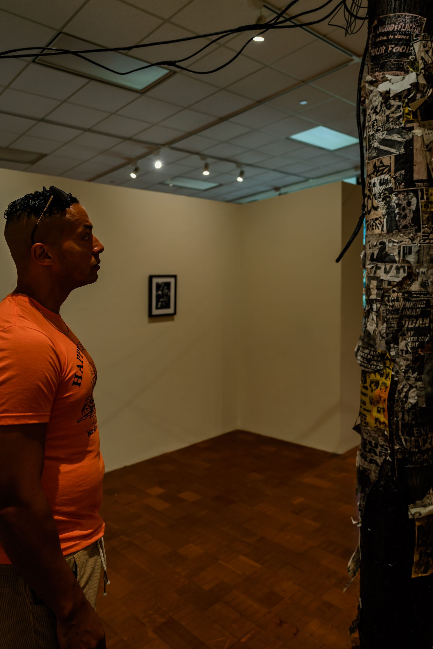 A man in an orange shirt examines a framed artwork near a pole covered in flyers in an art gallery.
