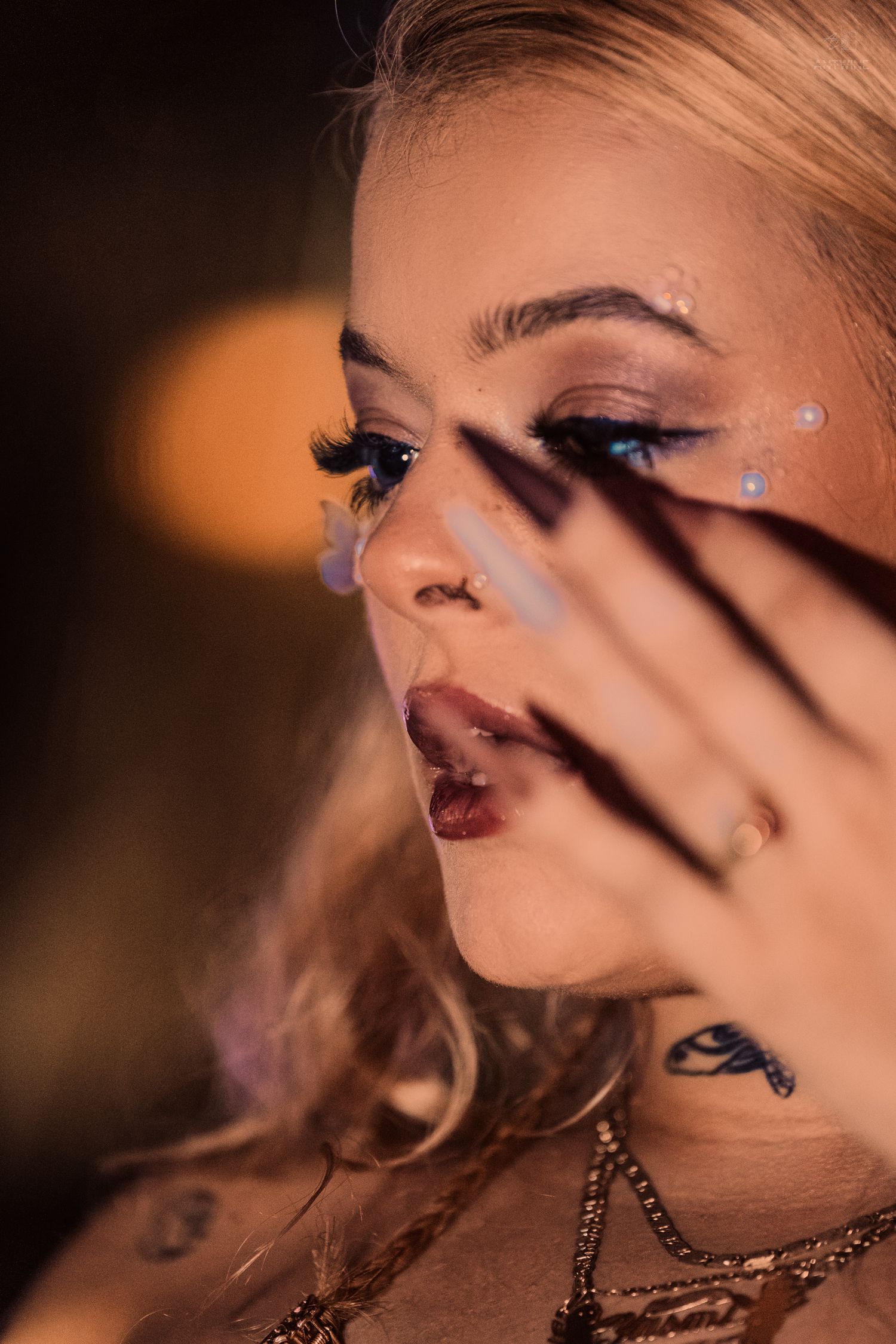 Close-up of blonde woman with blue eye makeup, glitter gems, long nails, and neck tattoos in warm light.
