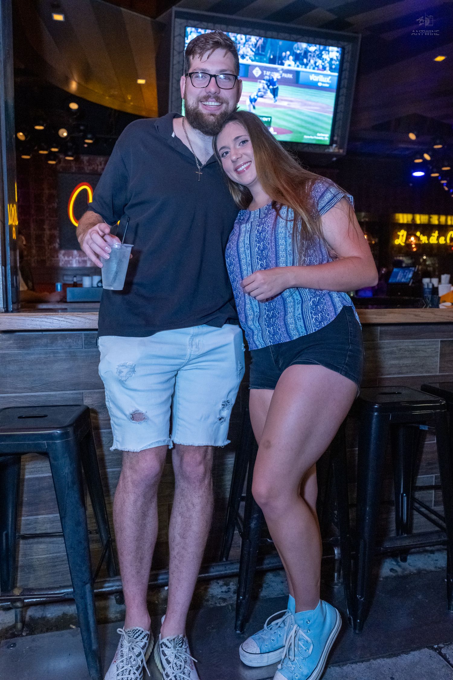 Smiling couple posing together at a bar with sports TV and bar stools nearby.