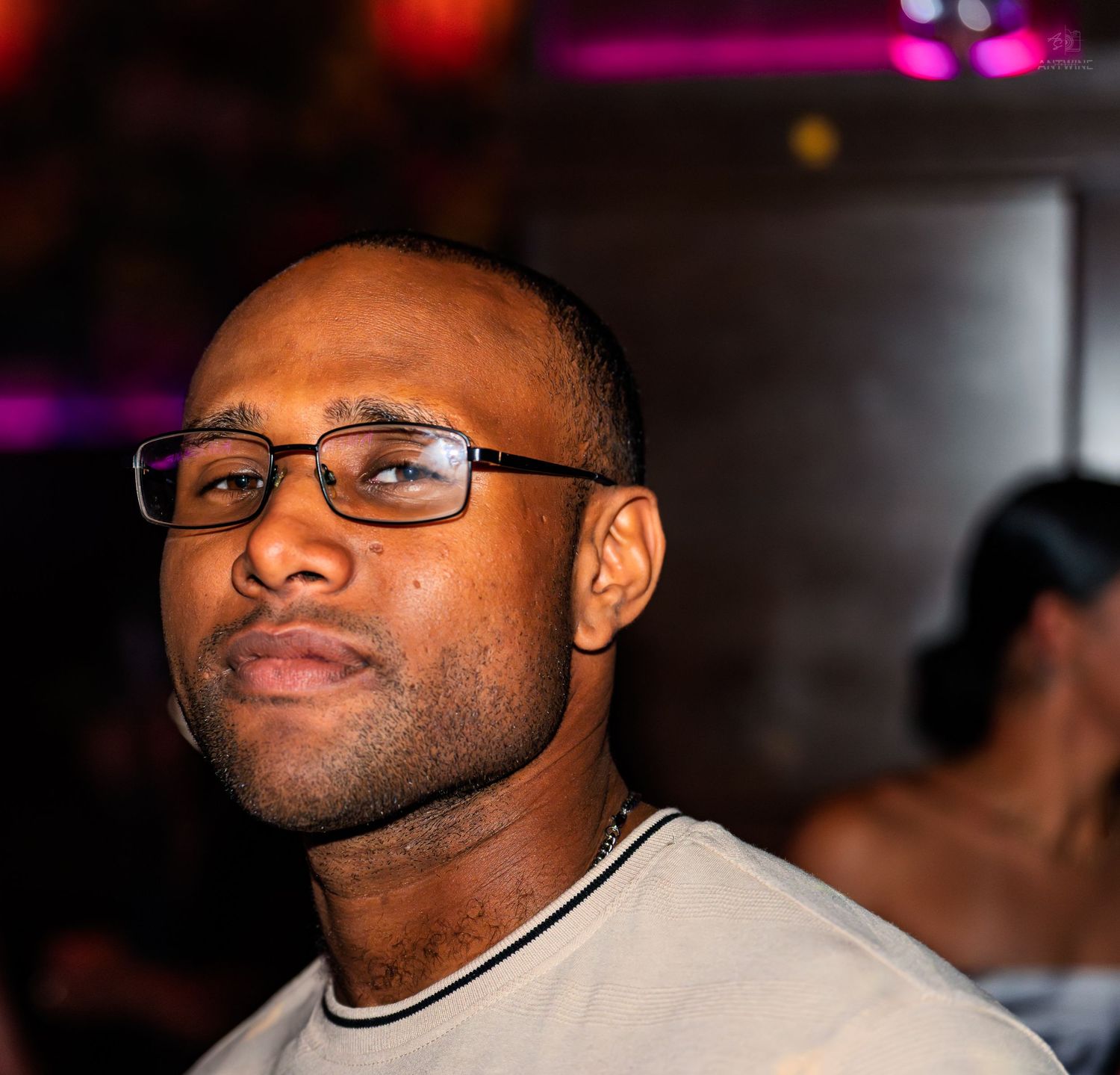 Man with glasses looking over his shoulder in a dimly lit nightclub with pink ambient lighting.