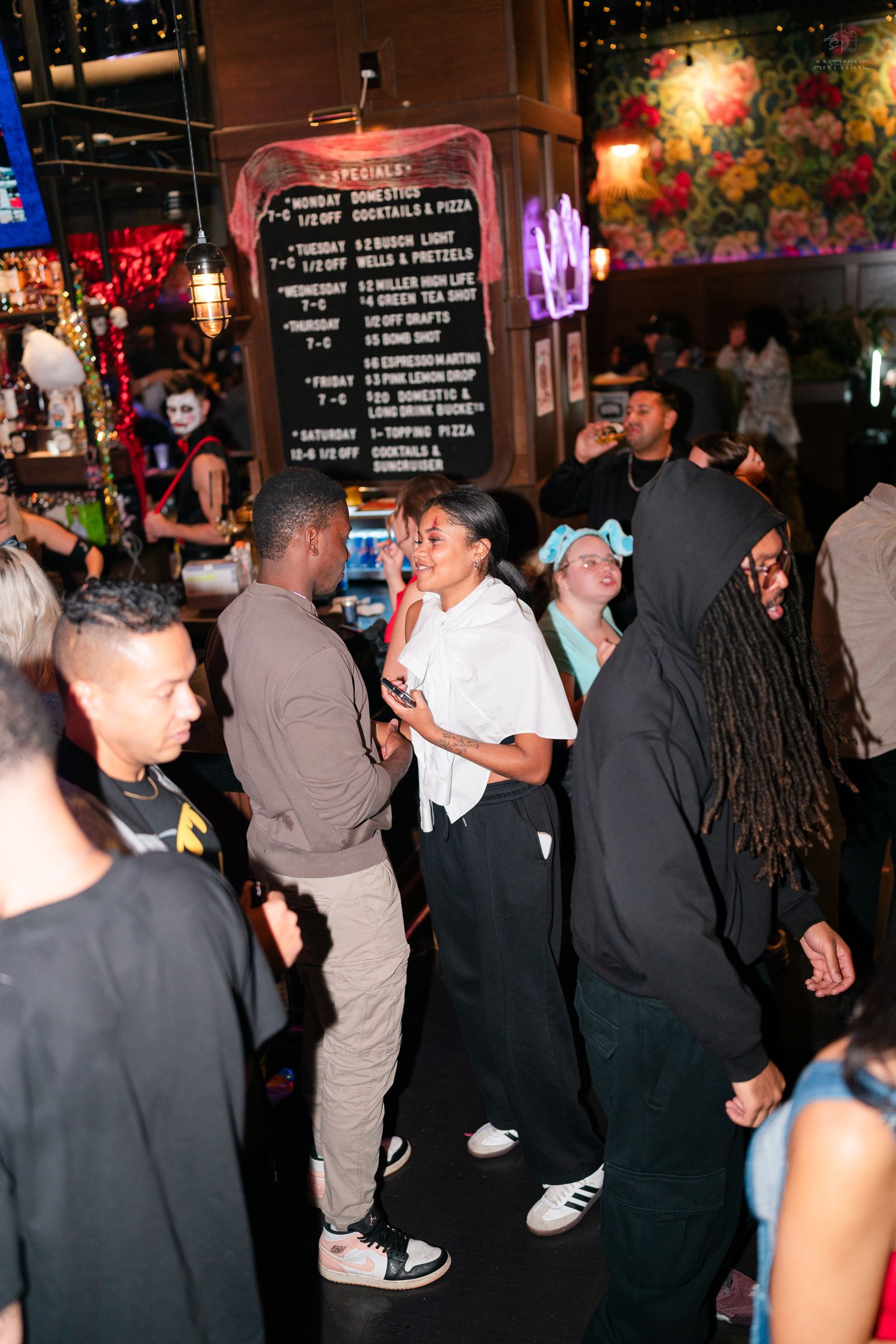 Diverse crowd socializing at a bar with a specials menu board in background.
