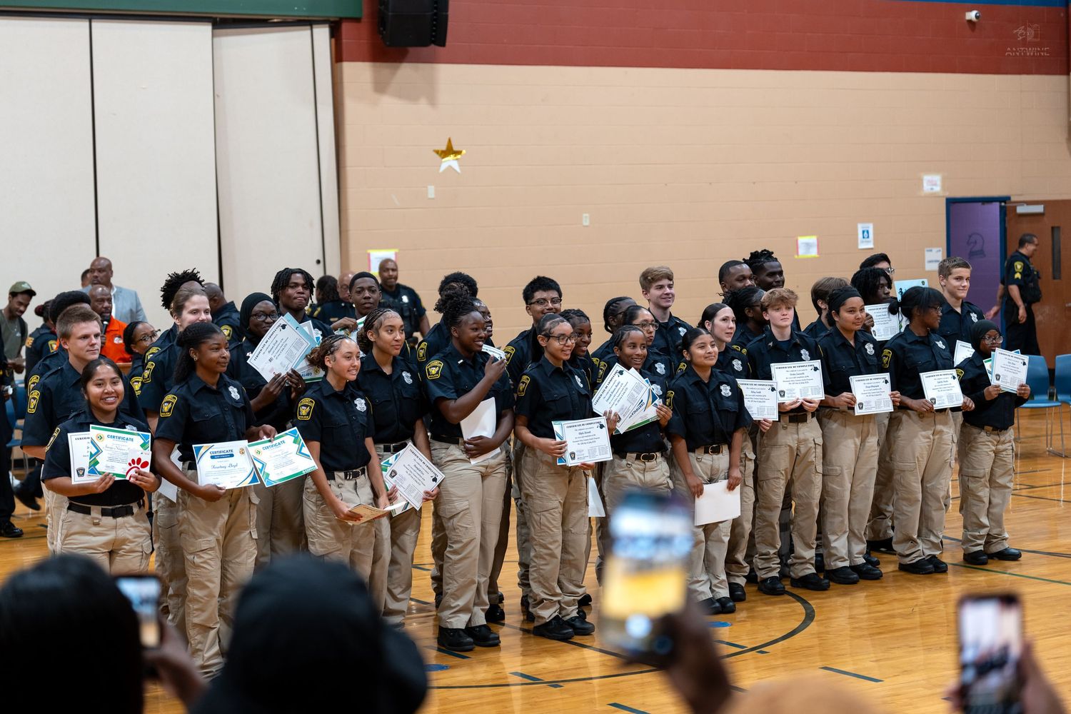 Students in uniform proudly hold certificates during an awards ceremony in a school gymnasium.