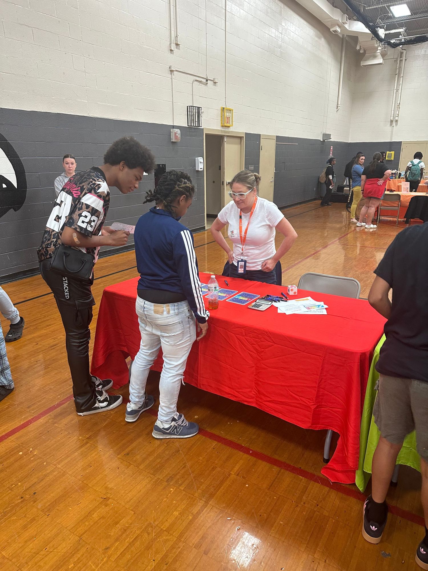 Two students consult with a representative at a red-draped table during an indoor school resource fair event.
