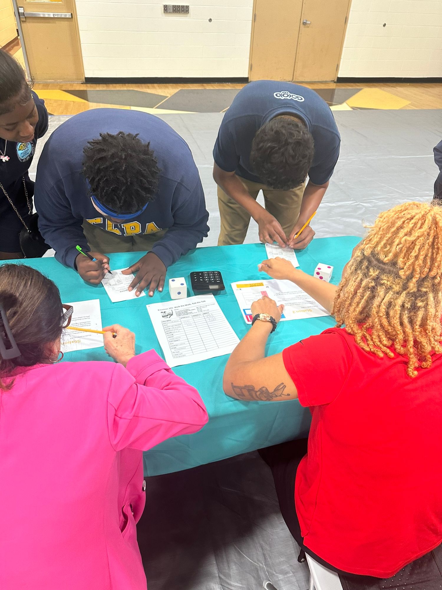Students gathered around a teal table completing worksheets together in a school gymnasium activity.