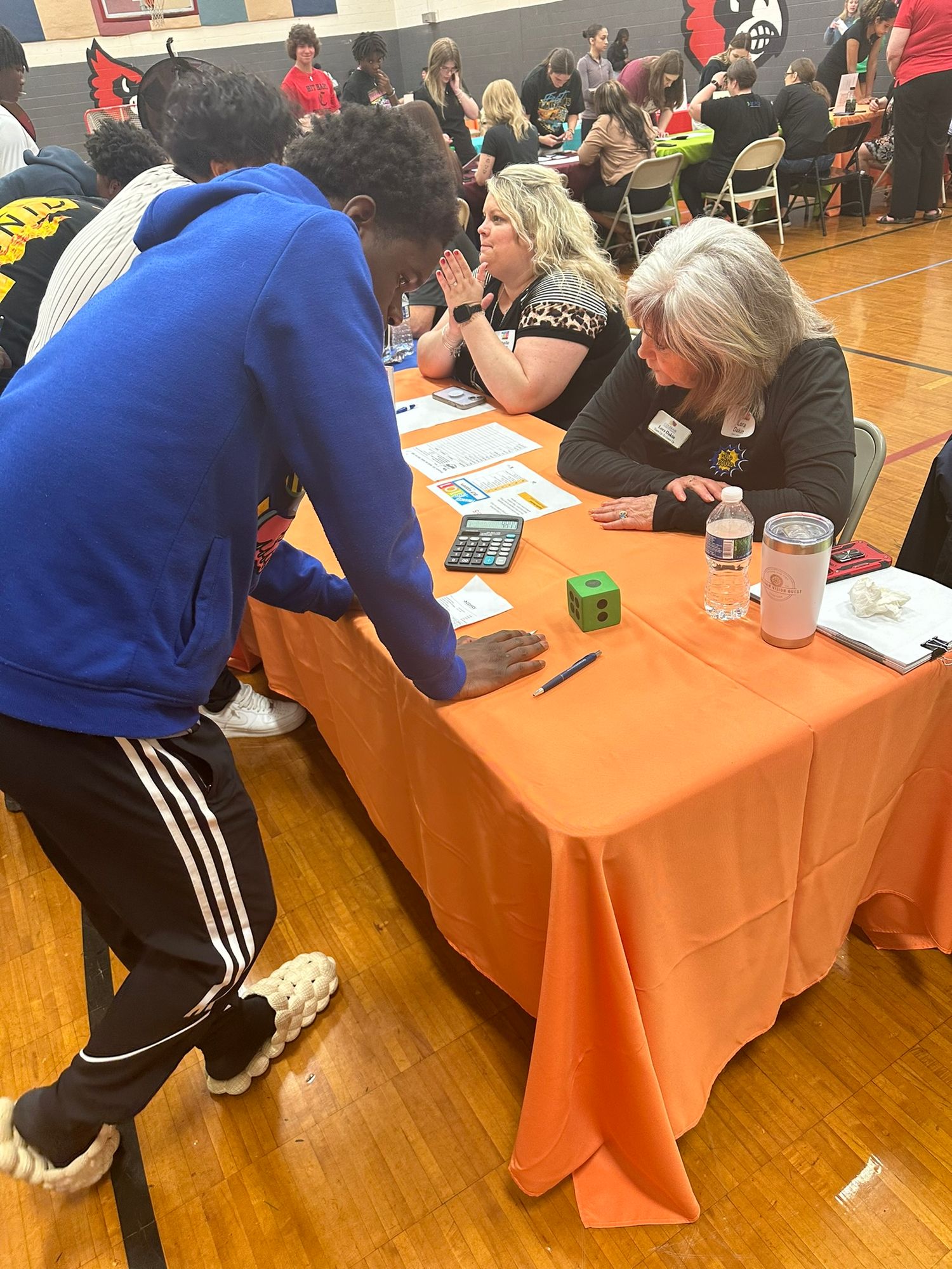 Students interact with adult advisors at orange-draped tables during a school career fair event in a gymnasium.