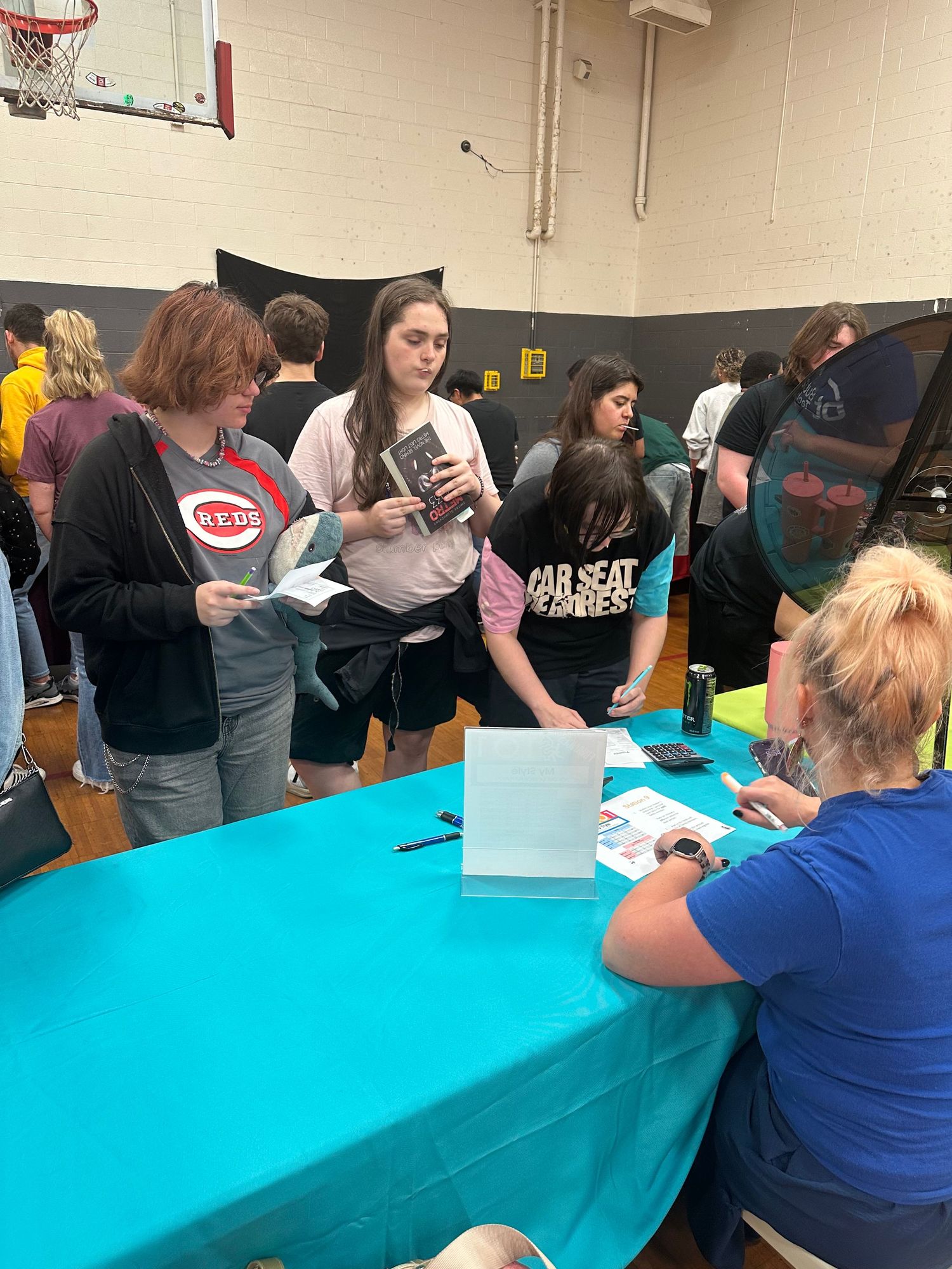 Students gather at a teal-covered table in a school gymnasium during a book signing or registration event.