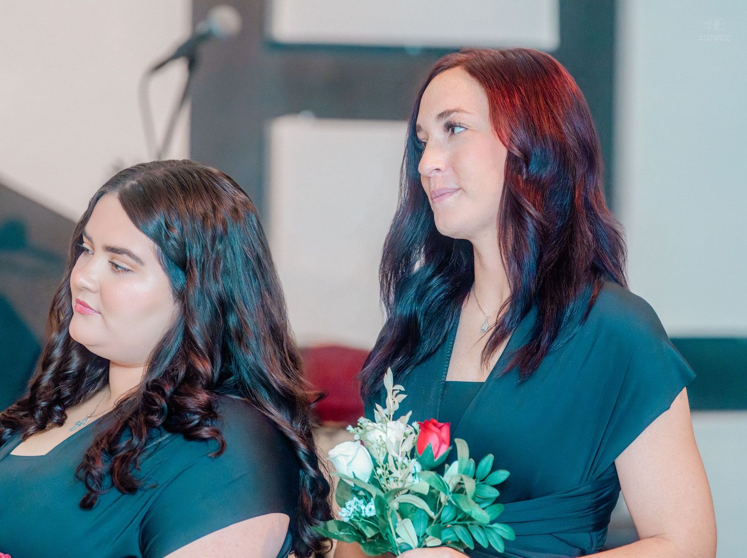 Two bridesmaids in dark green dresses, one holding a red rose bouquet, at a wedding.