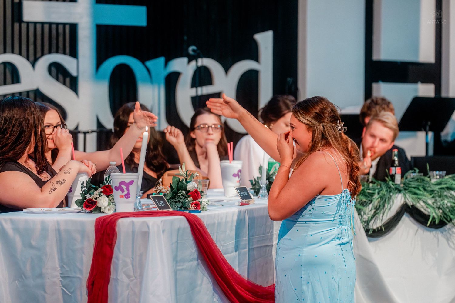 Bridesmaids in blue dresses raise glasses in a toast at a wedding reception with red table runners.