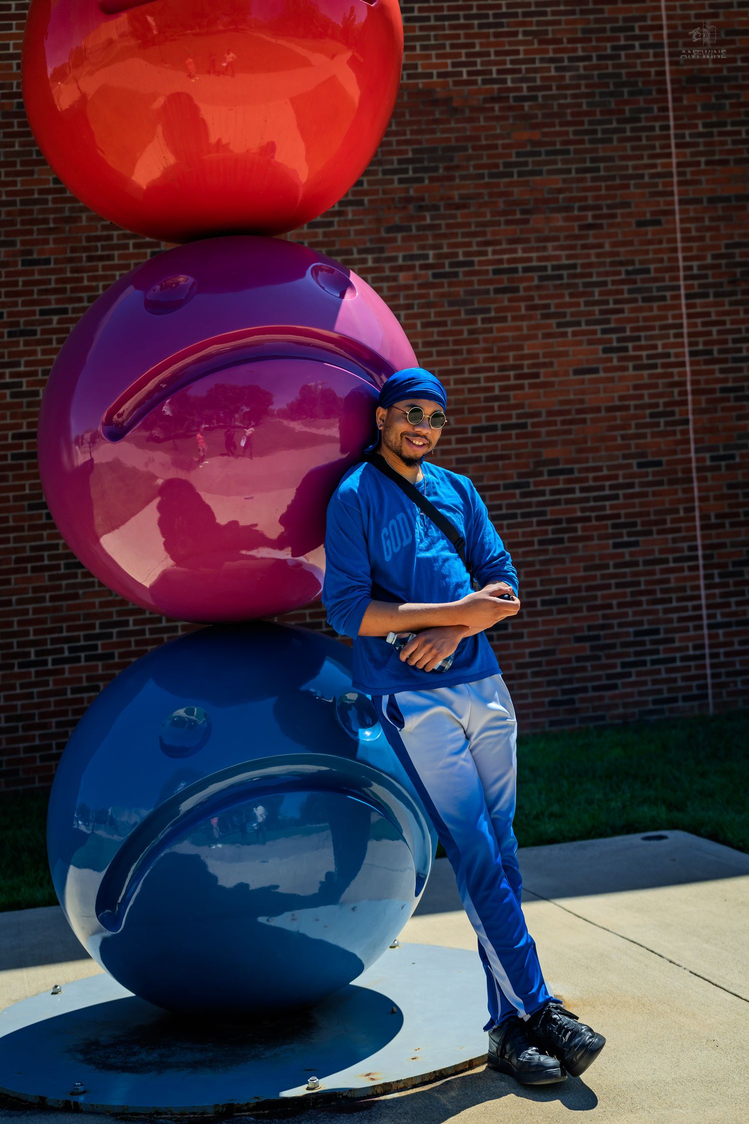 Man in blue clothes leaning against large colorful stacked ball sculpture by brick wall.