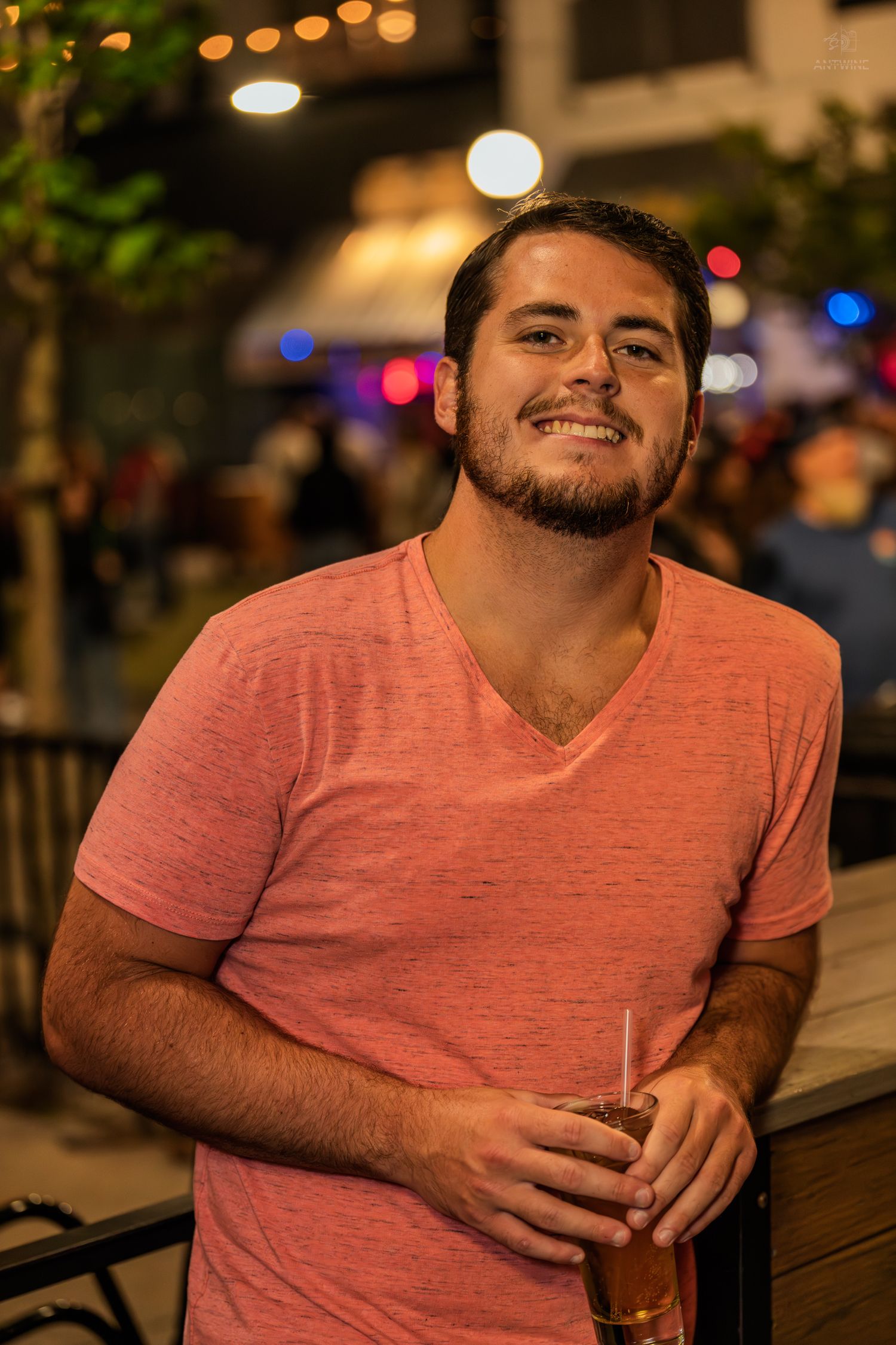 Young man in pink v-neck shirt smiling while holding a drink at an outdoor night bar event.