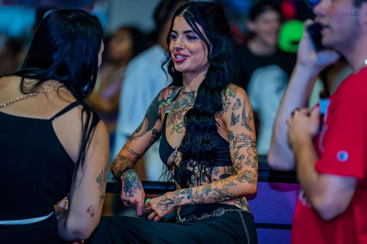 Heavily tattooed woman smiling at a lively nightclub event with colorful lighting.