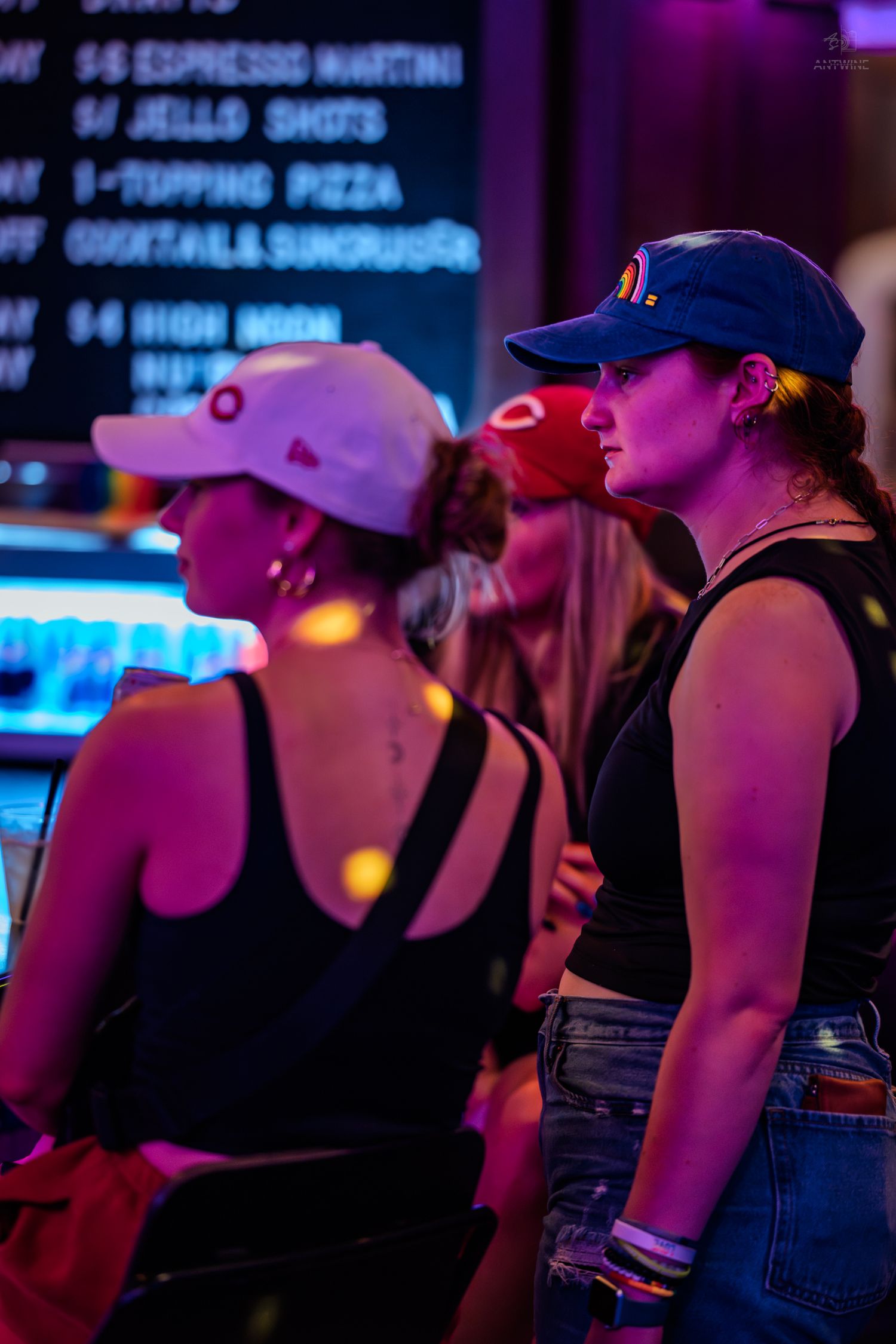 Two women wearing baseball caps standing in a colorful neon-lit bar at night.