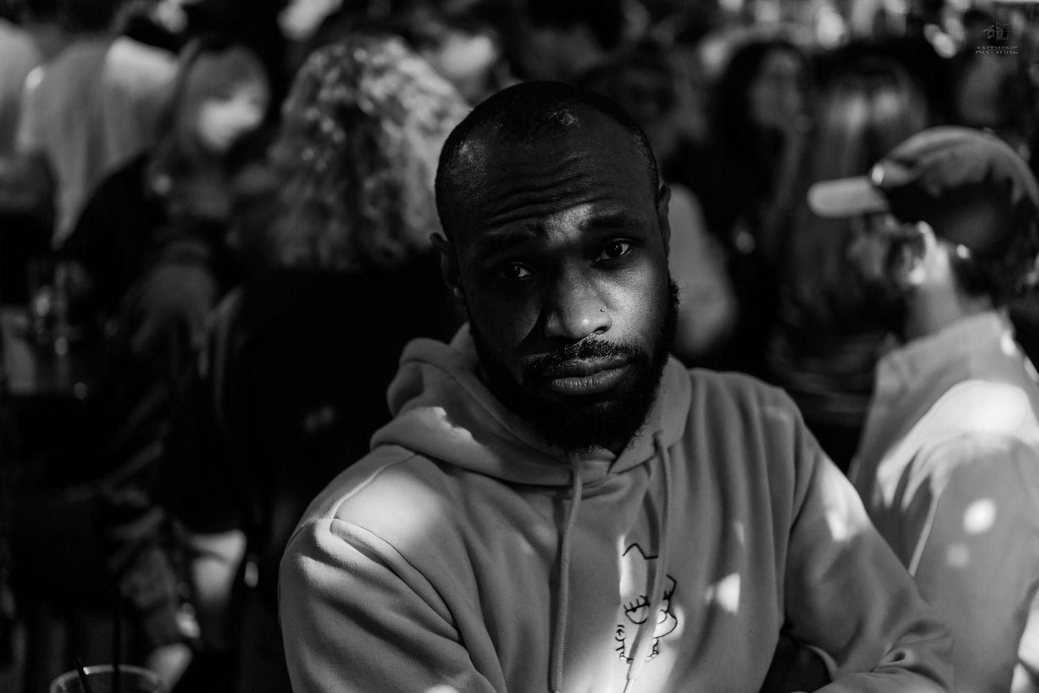Black and white close-up portrait of a bearded man in a hoodie at a crowded nighttime event.