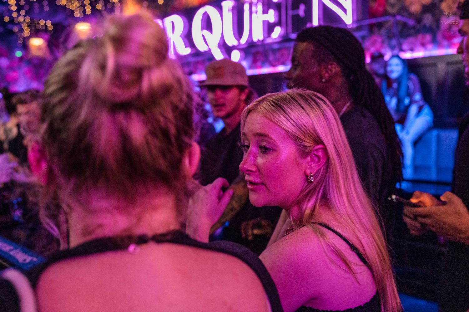 Two blonde women conversing at a neon-lit nightclub with purple and pink lighting.