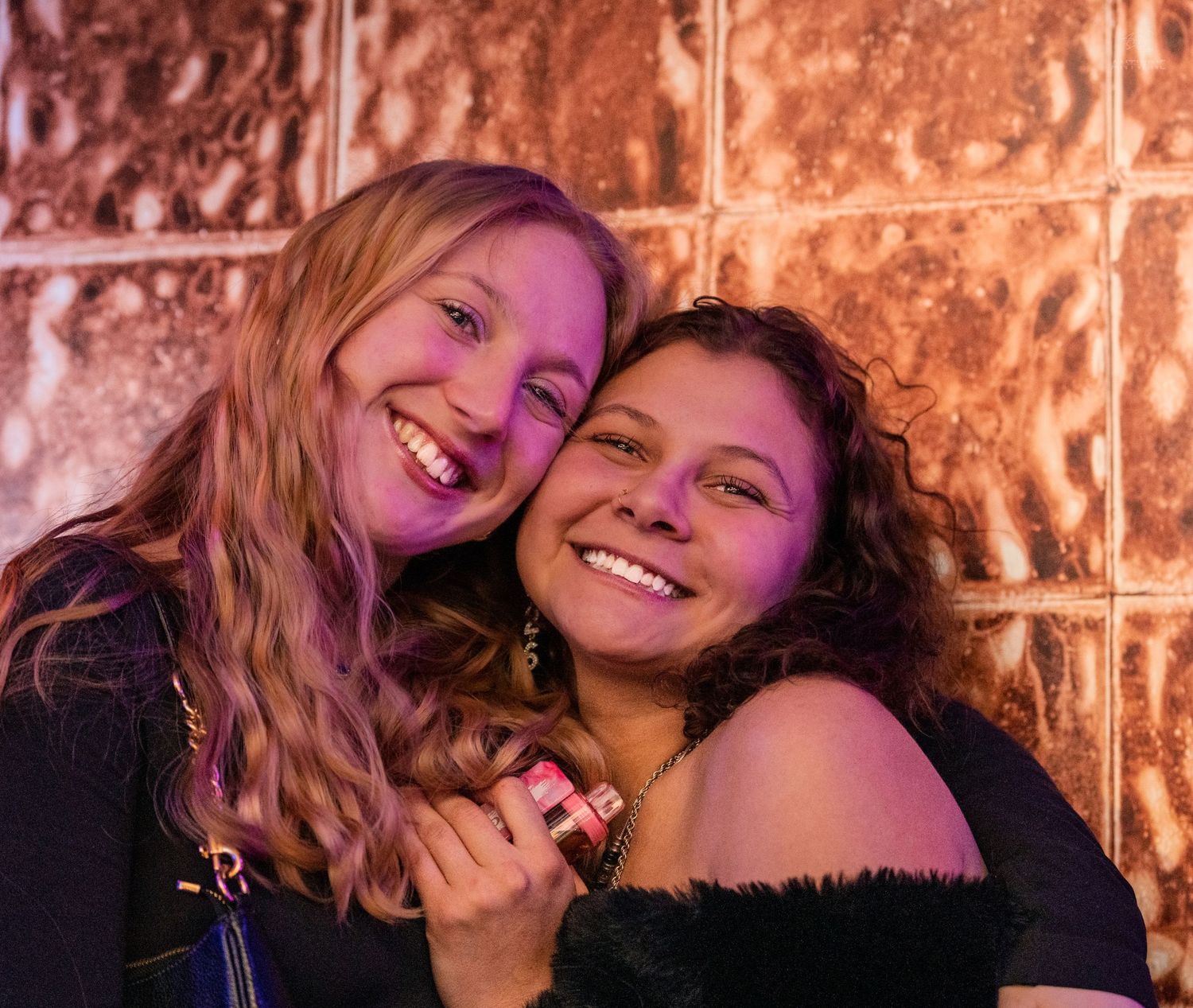 Two smiling women pose together against a glowing copper wall at a nightclub event.